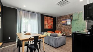 A modern living room with a dining area, a brick wall, a TV, and a kitchen area. - Stable #5 ~ Mini Golf, Fire Pits, Games, Walkable (Louisville)