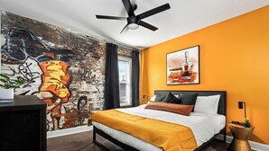 A modern bedroom with a brick wall mural, a bed with a mustard-colored throw, and a ceiling fan. - Stable #5 ~ Mini Golf, Fire Pits, Games, Walkable (Louisville)
