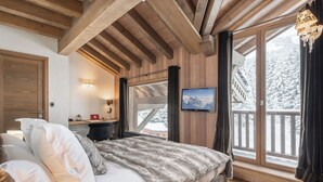 Ein gemütliches Schlafzimmer mit Holzbalkendecke, einem großen Bett mit weißer und grauer Bettwäsche, einem Fernseher an der Wand und Blick auf einen verschneiten Berg durch ein Fenster.