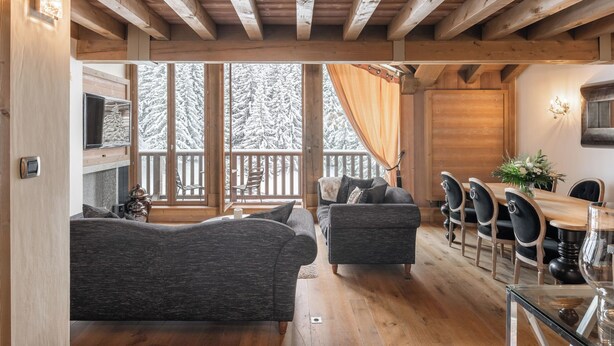 Ein gemütliches Blockhaus-Interieur mit Holzbalkendecke, einer Couch, einem Esstisch mit Stühlen und Blick auf schneebedeckte Bäume.