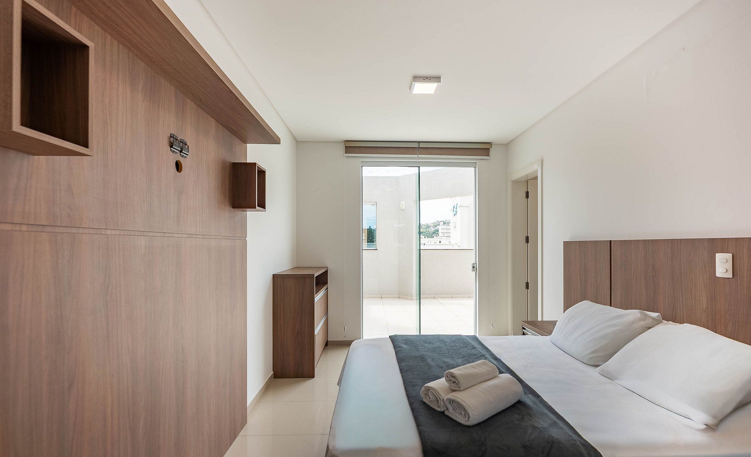 Un dormitorio moderno con una cama grande, paneles de madera, una mesita de noche pequeña y una puerta corrediza de vidrio que da a un balcón.