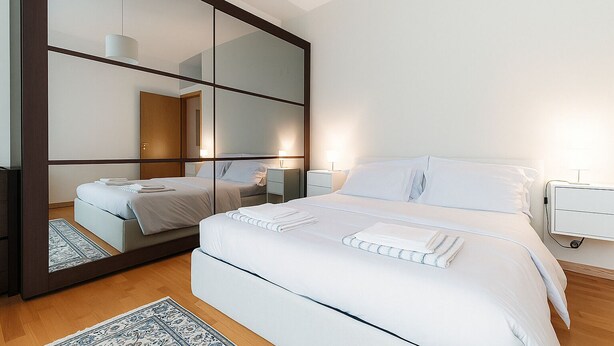 Een moderne slaapkamer met een grote spiegel, een bed met witte lakens, nachtkastjes en een kamerbreed tapijt met een patroon.