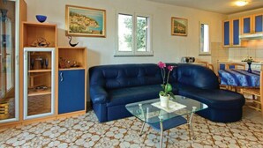 A living room with a blue sofa, a glass coffee table, and a kitchen area with wooden cabinets. - Holiday Home Brsec-Two Bedroom Holiday Home with Sea View (Brsec)