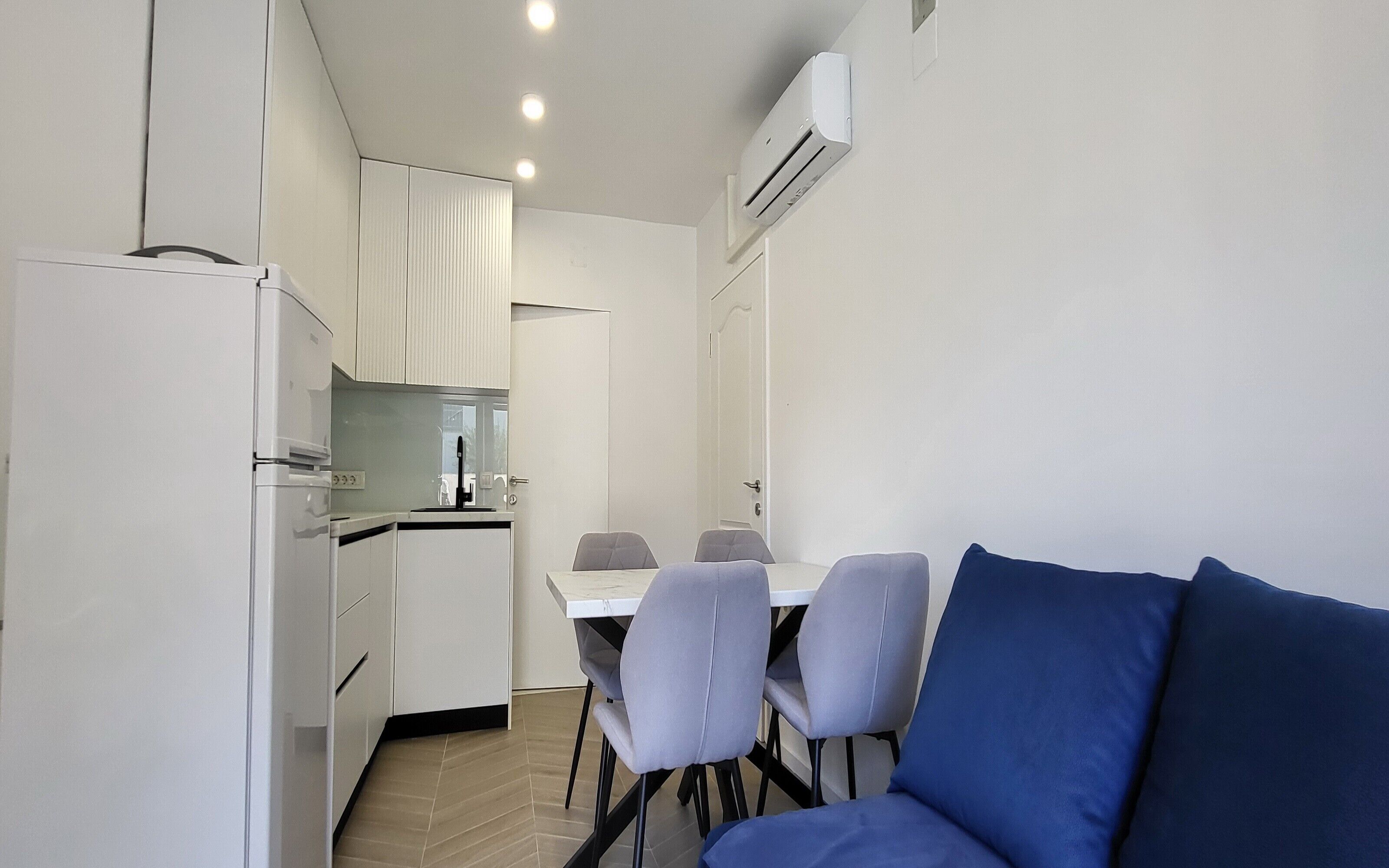 A compact kitchen with white cabinets, a dining table with four chairs, and a blue sofa.