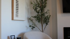 Ein modernes Wohnzimmer mit einem runden, hellen Sessel, einem kleinen Holztisch mit Bilderrahmen und Tasse, einer großen Topfpflanze und einem flach an der Wand befestigten Fernseher.