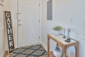A hallway with a door, a welcome sign, a small table with decorative items, and a patterned rug. - Updated Large 2BR in Azalea Lakes, Pools, PondView (Myrtle Beach)