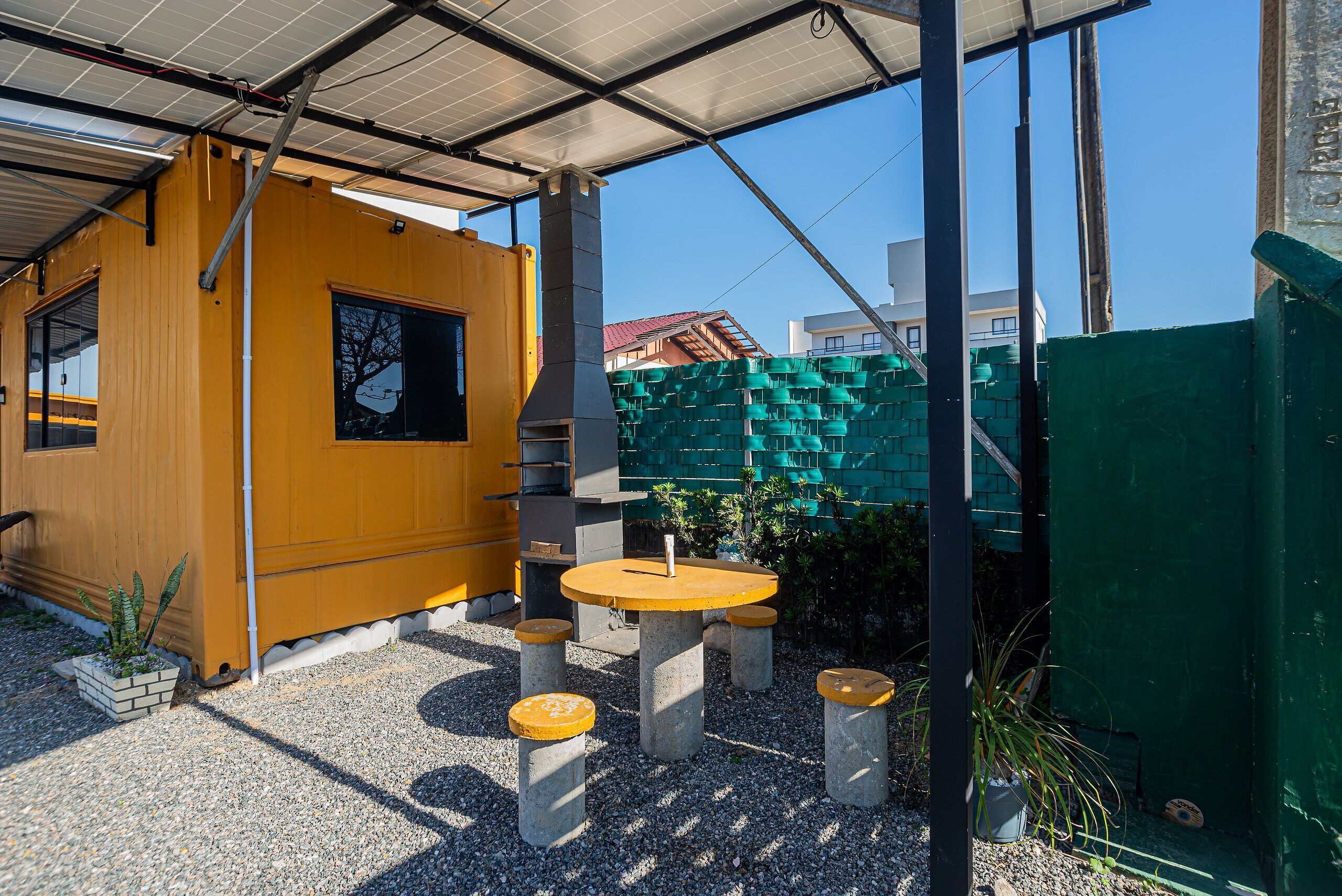 Un edificio amarillo con un espacio al aire libre cubierto, que incluye una mesa y taburetes, rodeado de grava y plantas en macetas.