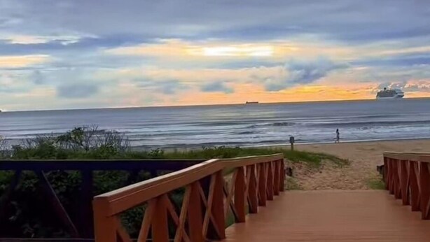 A wooden boardwalk leading to a beach with a cloudy sky and a ship in the distance. - Container Beira Mar Solar Analuz (Navegantes)
