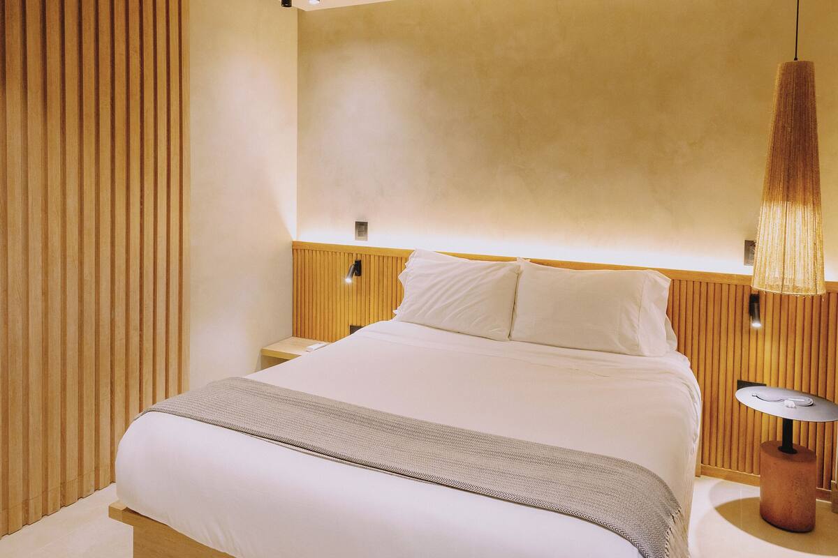 A modern bedroom with a wooden headboard, a bedside table with a lamp, and a large bed with white linens.