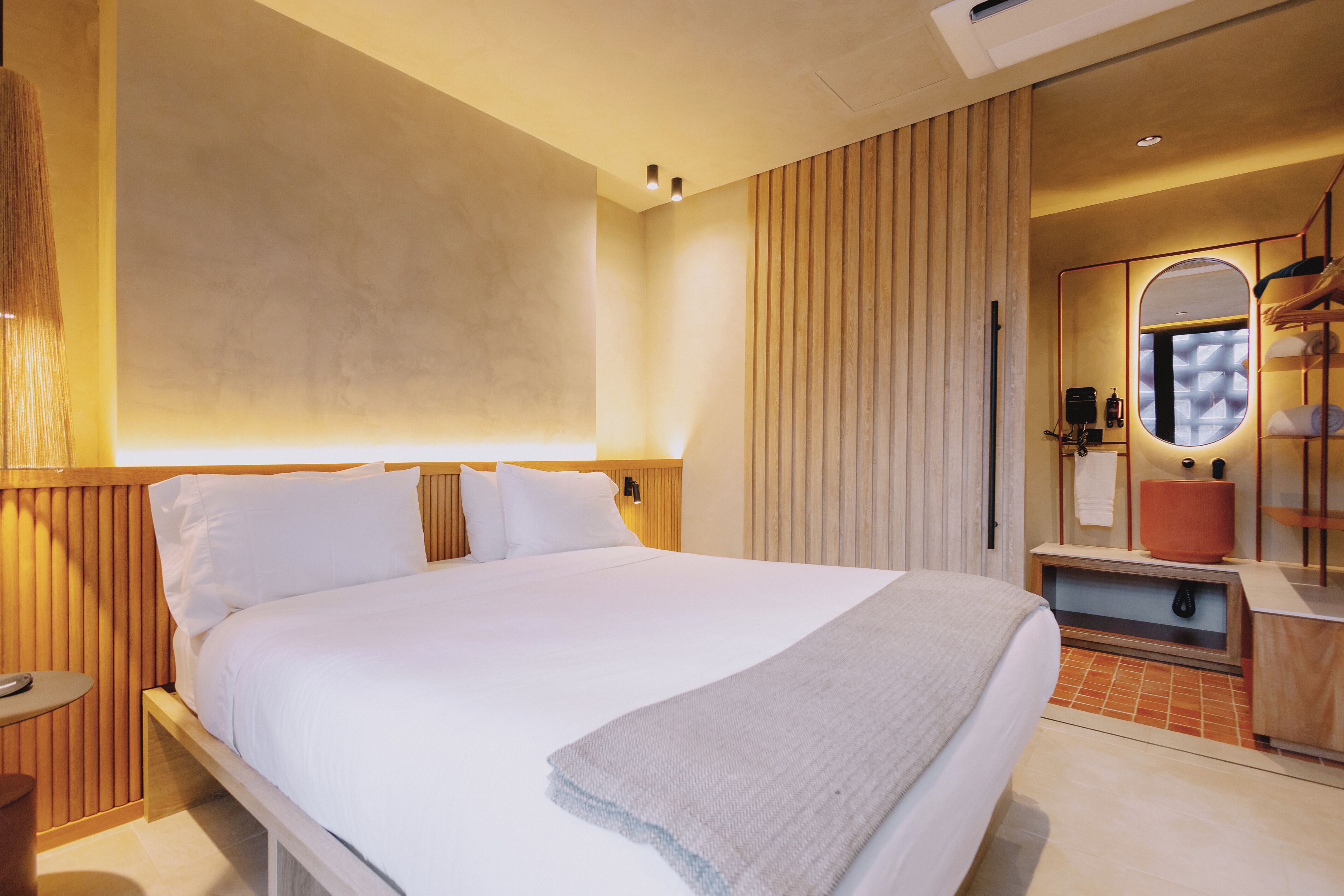 A hotel room with a large bed, a wooden headboard, a bedside table, a mirror, a towel rack, and a small bathroom area with a sink and mirror.