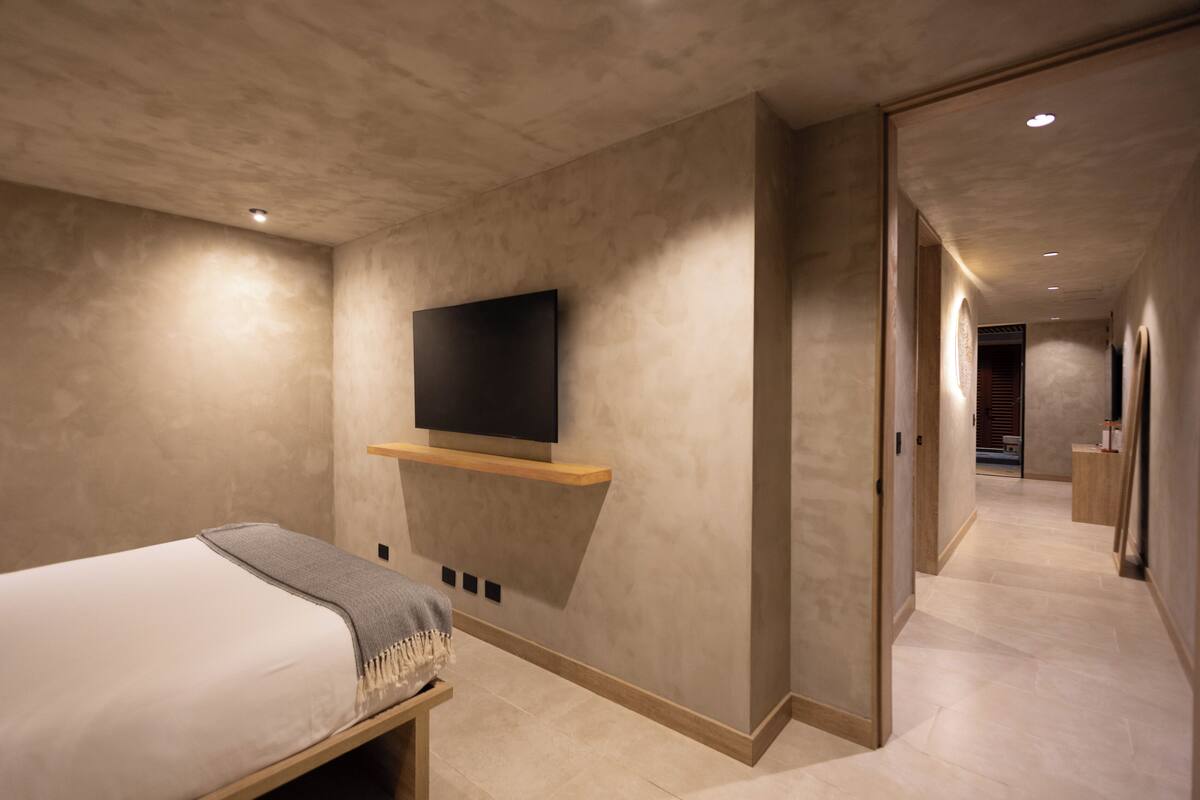 A modern hotel room with a bed, a mounted TV, and a hallway leading to a bathroom.