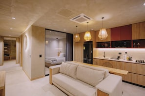 A modern hotel room with a sofa, a dining table, a kitchen area with a microwave, and a separate bedroom with a bed.