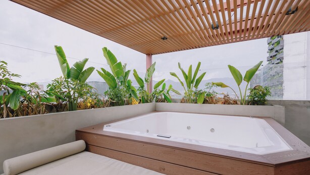 A hot tub under a wooden pergola with a view of plants and a building.