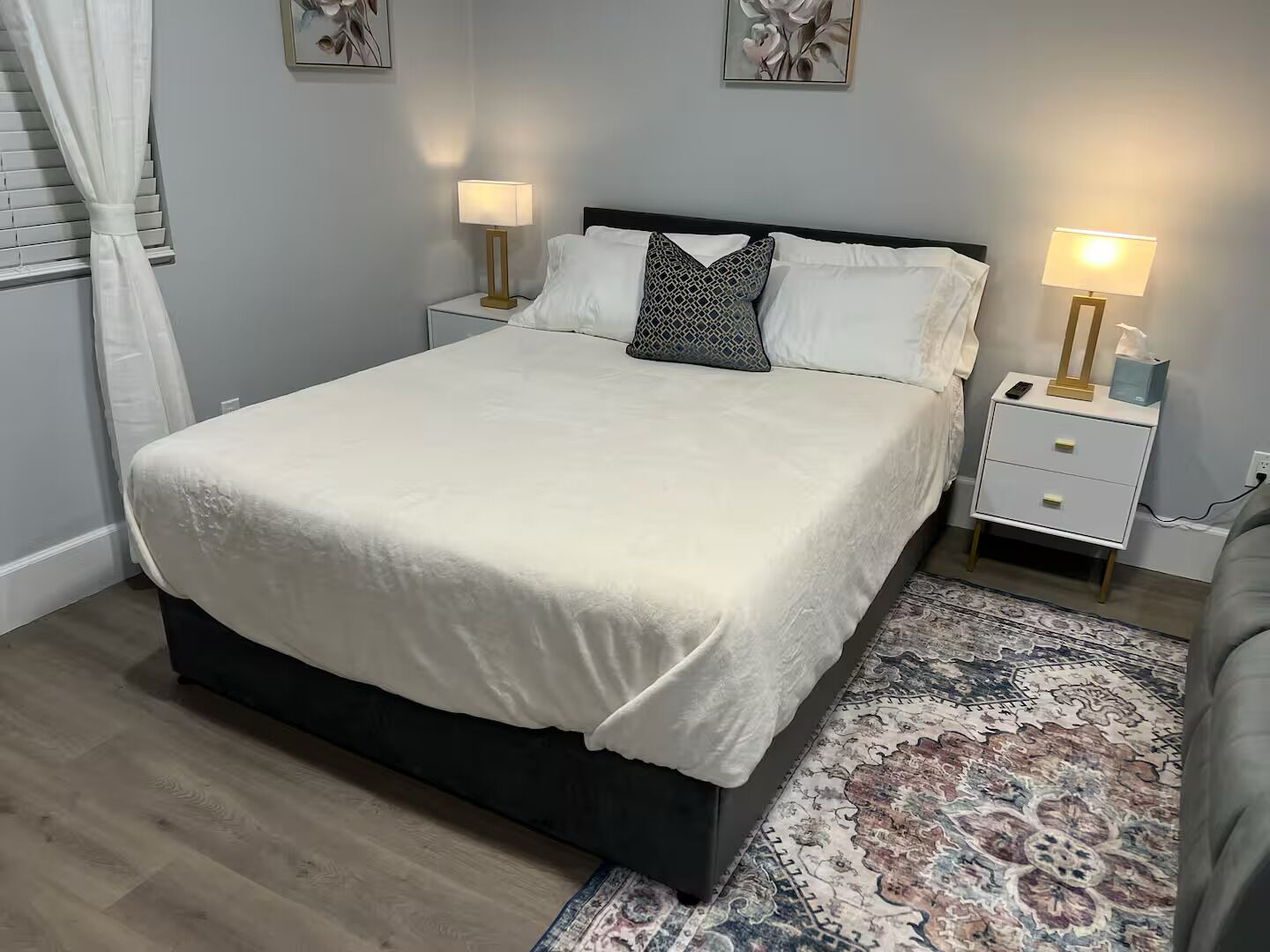 A neatly made bed with white linens, two bedside tables with lamps, a patterned rug, and artwork on the walls.