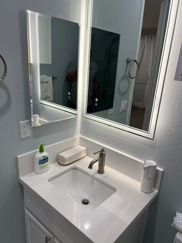 Un baño con lavamanos blanco, espejo, una botella de jabón Cetaphil y una pila de toallas.