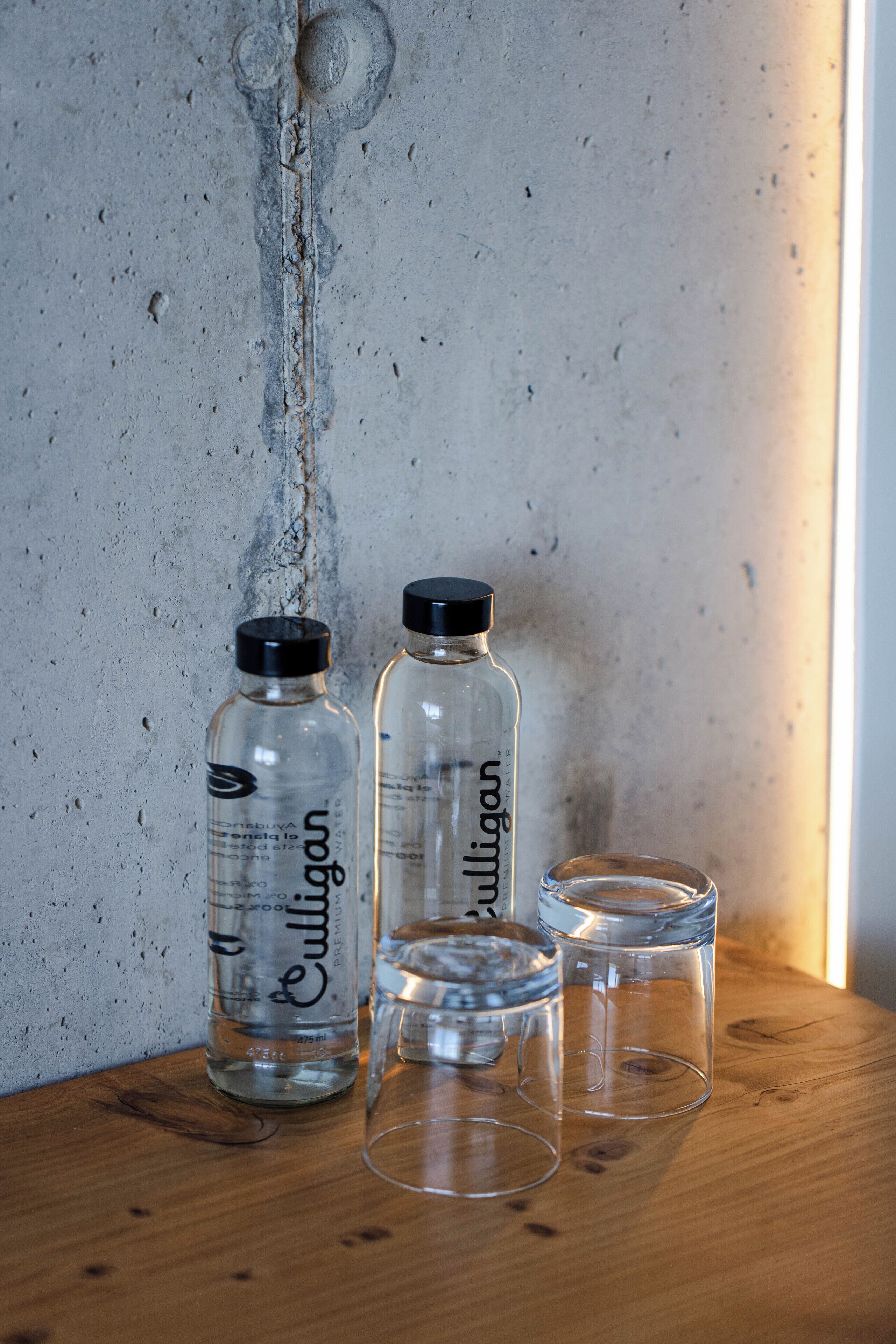 Two clear glass bottles with black caps and a wooden surface with three empty glass cups.