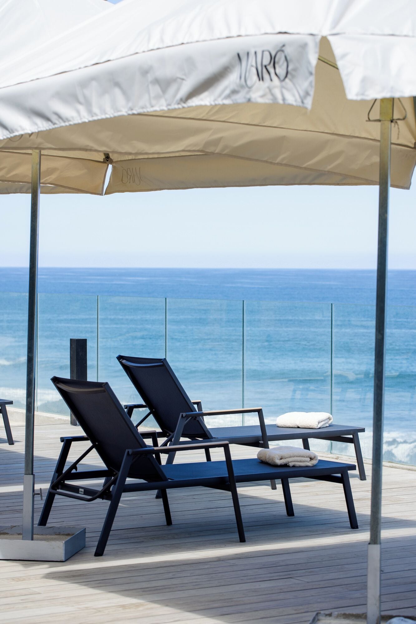 A beachside lounge with two lounge chairs and a table under a canopy, overlooking the ocean.