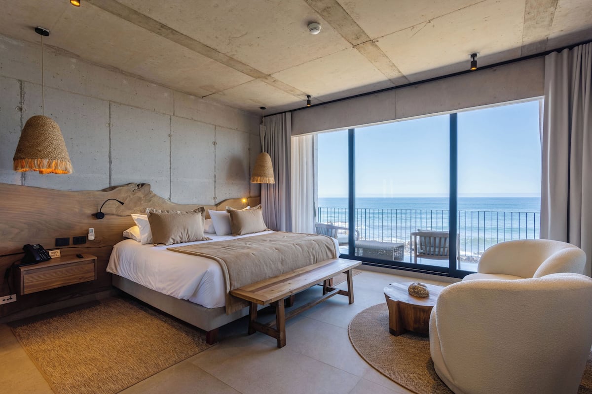 Ett modernt hotellrum med en stor säng, en träbänk, en vit fåtölj och utsikt över havet.