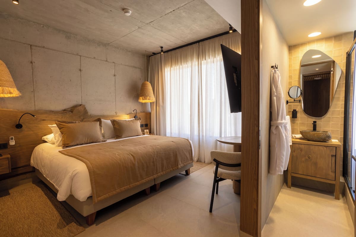 Ett modernt hotellrum med en stor säng, ett nattduksbord, en stol, ett litet runt bord, en spegel och ett badrum med handfat och handduksställ.