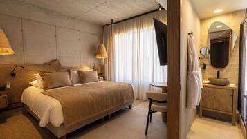 Una habitación de hotel moderna con una cama grande, una mesita de noche, una silla, una pequeña mesa redonda, un espejo y un baño con lavabo y toallero.