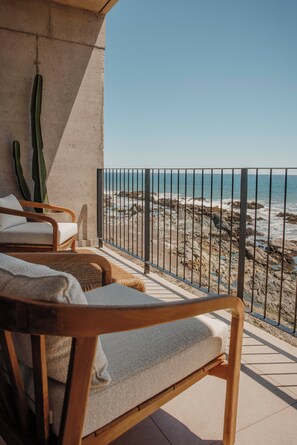 A balcony with two wooden chairs and a woven chair, overlooking a rocky beach with the ocean.