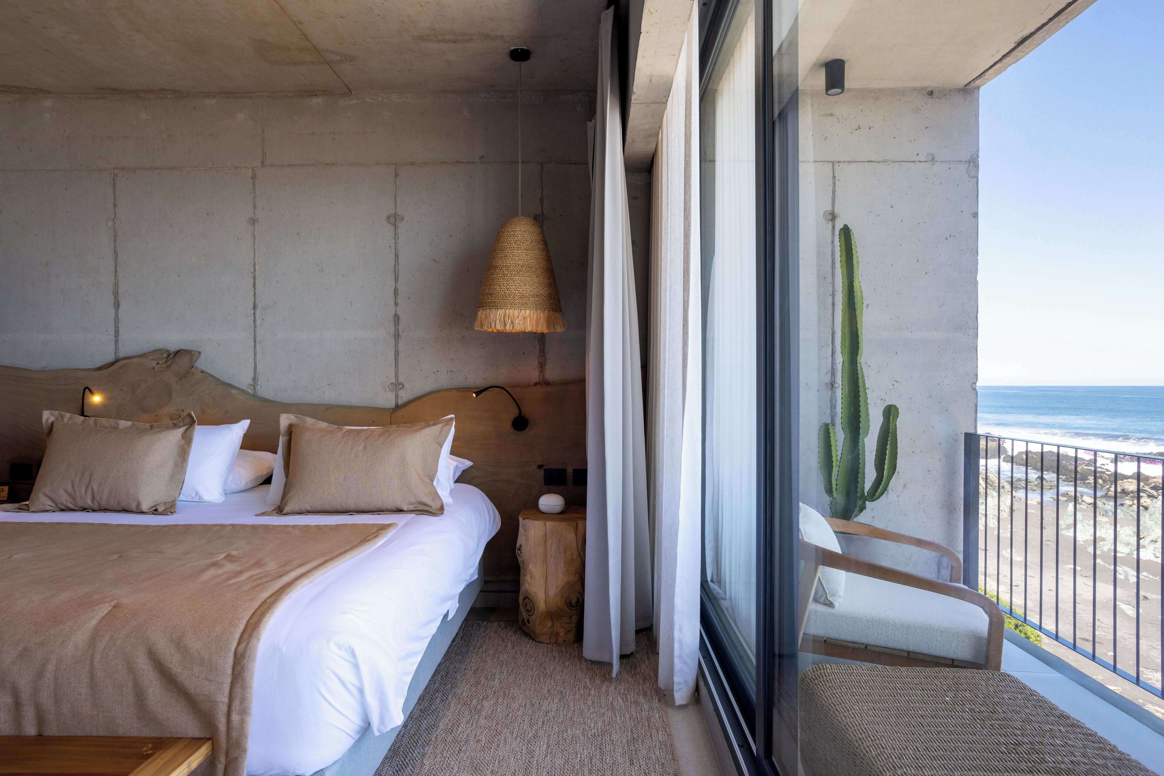 Une chambre avec un grand lit, une table de chevet, une lampe, un tabouret en bois, une chaise et une vue sur la plage et l’océan.