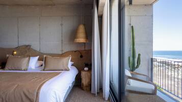 Une chambre avec un grand lit, une table de chevet, une lampe, un tabouret en bois, une chaise et une vue sur la plage et l’océan.