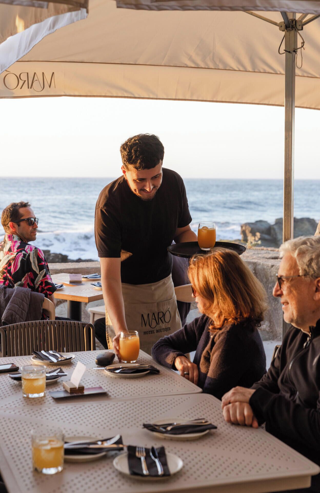 Un restaurant al mar amb taules preparades per dinar, un cambrer servint begudes i una vista a l'oceà.