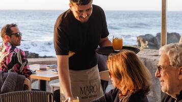 Un restaurant al mar amb taules preparades per dinar, un cambrer servint begudes i una vista a l'oceà.