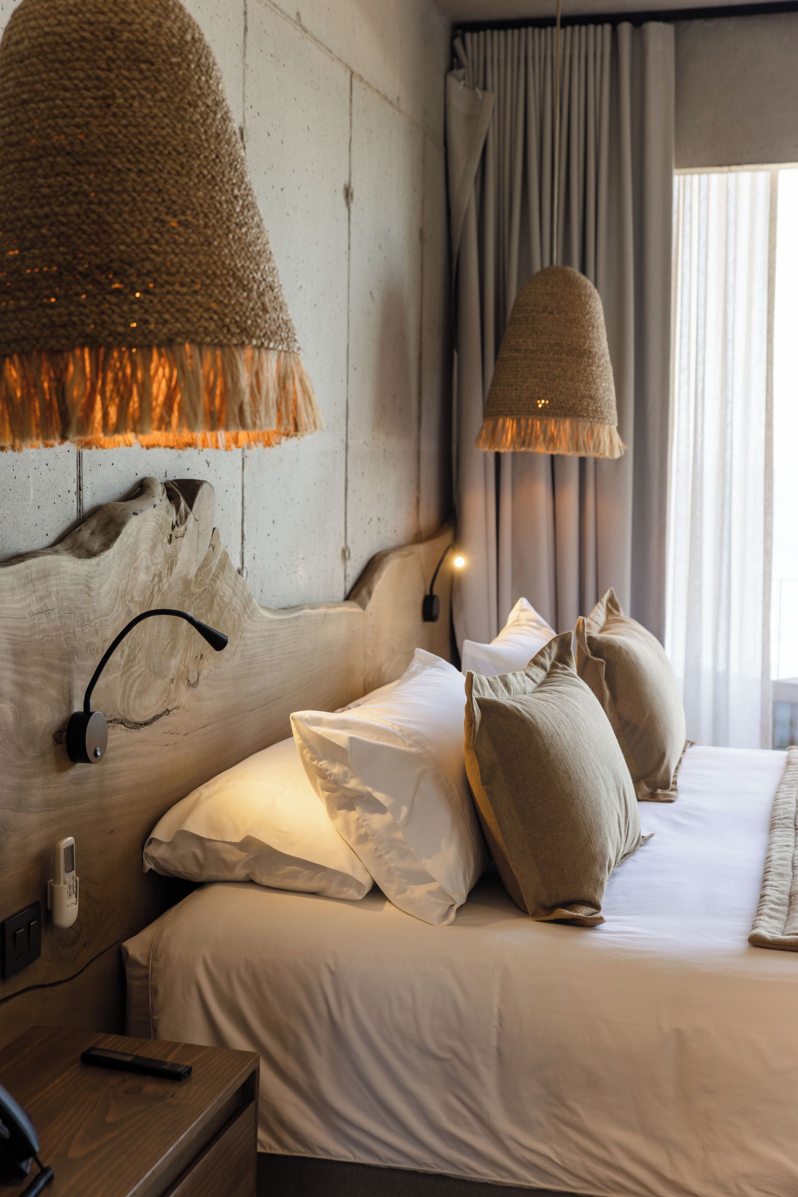 A hotel room with a wooden headboard, a bedside table with a lamp, a bed with beige bedding, and a view of the outdoors through curtains.