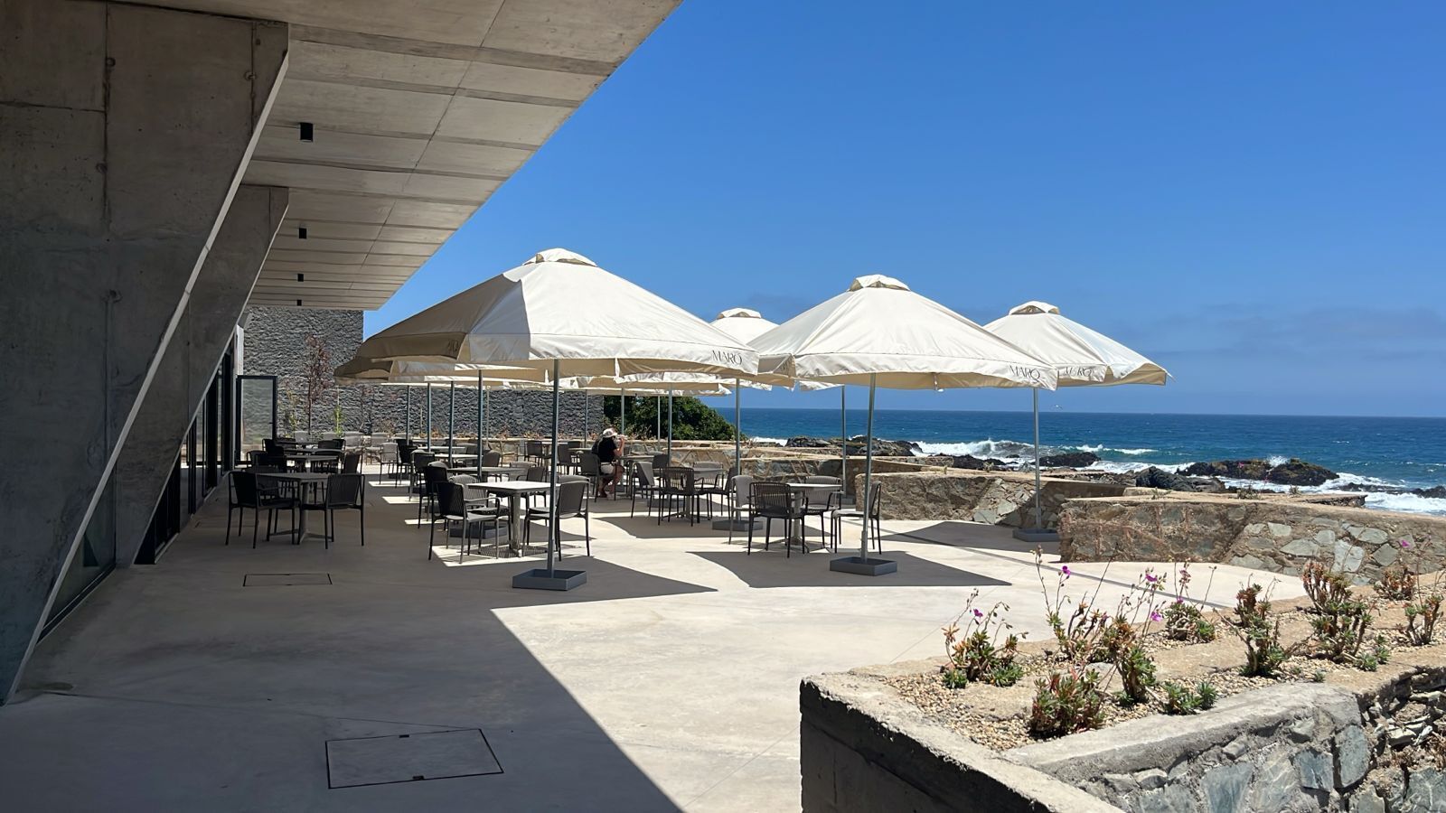 An outdoor seating area with tables and chairs under umbrellas, overlooking the ocean.
