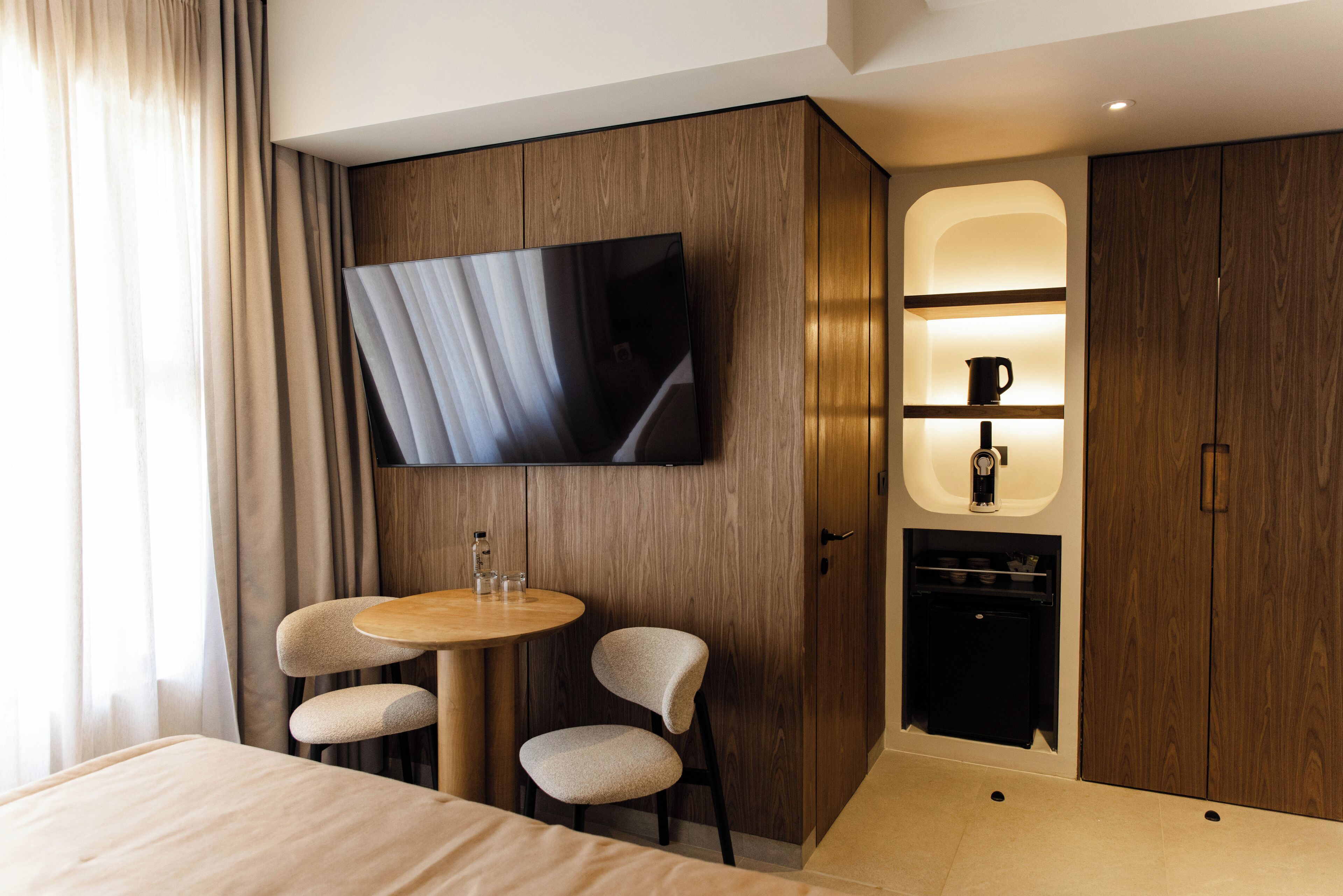 A modern hotel room with a bed, a TV mounted on the wall, a small table with chairs, and a built-in mini-fridge.