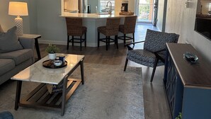 Ein modernes Wohnzimmer mit einer Couch, einem Couchtisch und einem gemusterten Teppich. Es gibt eine Küchenzeile mit einer großen Kochinsel und Barhocker.