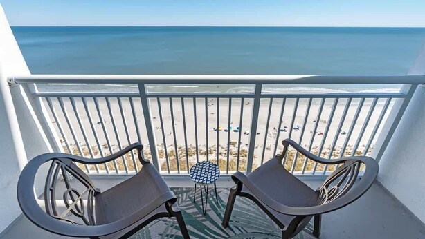 A balcony with two chairs and a small table, overlooking a beach with the ocean in the background. - 1BR Camelot Resort, Direct Oceanfront Views,Pools! (Myrtle Beach)