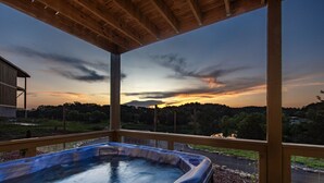A hot tub under a wooden pergola with a view of a sunset over a forested landscape.