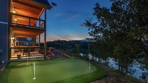 A house with a covered patio and a putting green by a lake.