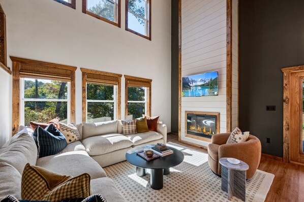 A spacious living room with a large sectional sofa, a round coffee table, and a fireplace. There are large windows with wooden frames, and a television mounted on the wall.