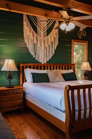 Uma cama de madeira com cabeceira, duas mesas de cabeceira com abajures, um painel de macramé na parede e um ventilador de teto.