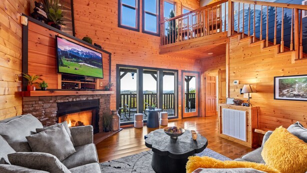 A cozy cabin interior with a fireplace, comfortable seating, a TV, and a balcony with a scenic view. - The Real McCoy | Epic Views, Hot Tub, Arcade! (Sevierville)