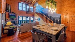 A spacious living area with a fireplace, a dining table set for four, and a staircase leading to a loft. - The Real McCoy | Epic Views, Hot Tub, Arcade! (Sevierville)