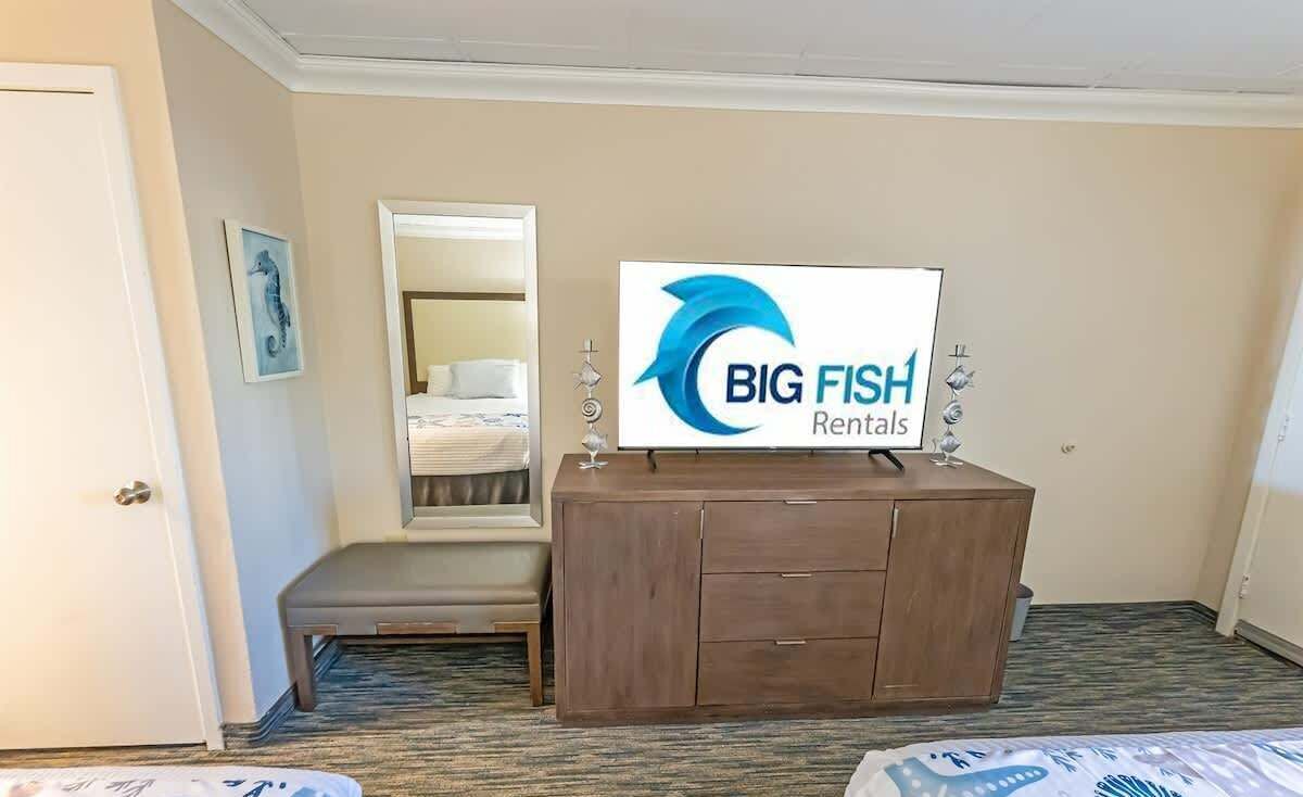 Une chambre d’hôtel avec une commode en bois, un lit, un banc et un miroir. Une pancarte sur la commode indique : « BIG FISH Rentals ».