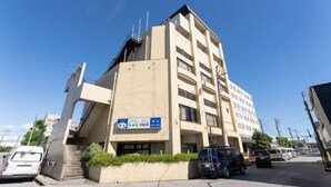 Un immeuble à plusieurs étages, doté d’une façade beige, d’une enseigne bleue avec du texte blanc, et d’un espace de stationnement où se trouvent plusieurs véhicules.