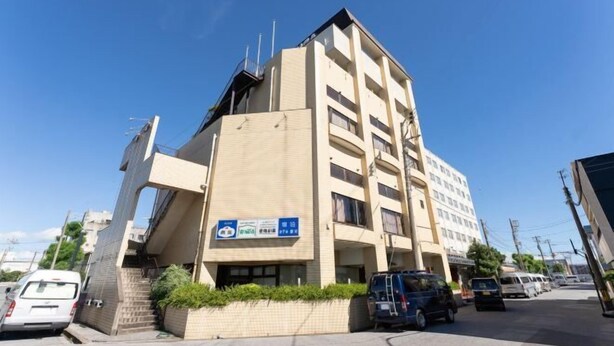 Un immeuble à plusieurs étages, doté d’une façade beige, d’une enseigne bleue avec du texte blanc, et d’un espace de stationnement où se trouvent plusieurs véhicules.