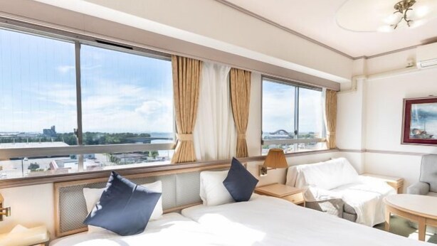 Une chambre d’hôtel avec un grand lit, deux tables de chevet, une petite table et une vue sur le paysage urbain grâce à de grandes fenêtres.