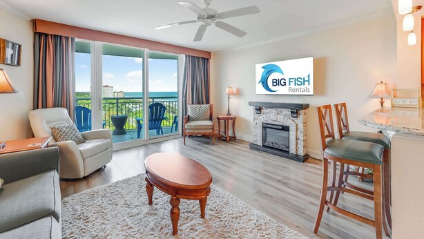 A spacious living room with a fireplace, a ceiling fan, and a large TV screen displaying 'BIG FISH Rentals'. - New 2BR at Horizon at 77th with Views,Nice Pools! (Myrtle Beach)