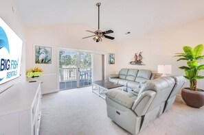A spacious living room with a grey sofa, a coffee table, a ceiling fan, and a large window with a view of the outdoors. - NEW, Large 2 BR at Magnolia Place,Golf Course View (Myrtle Beach)