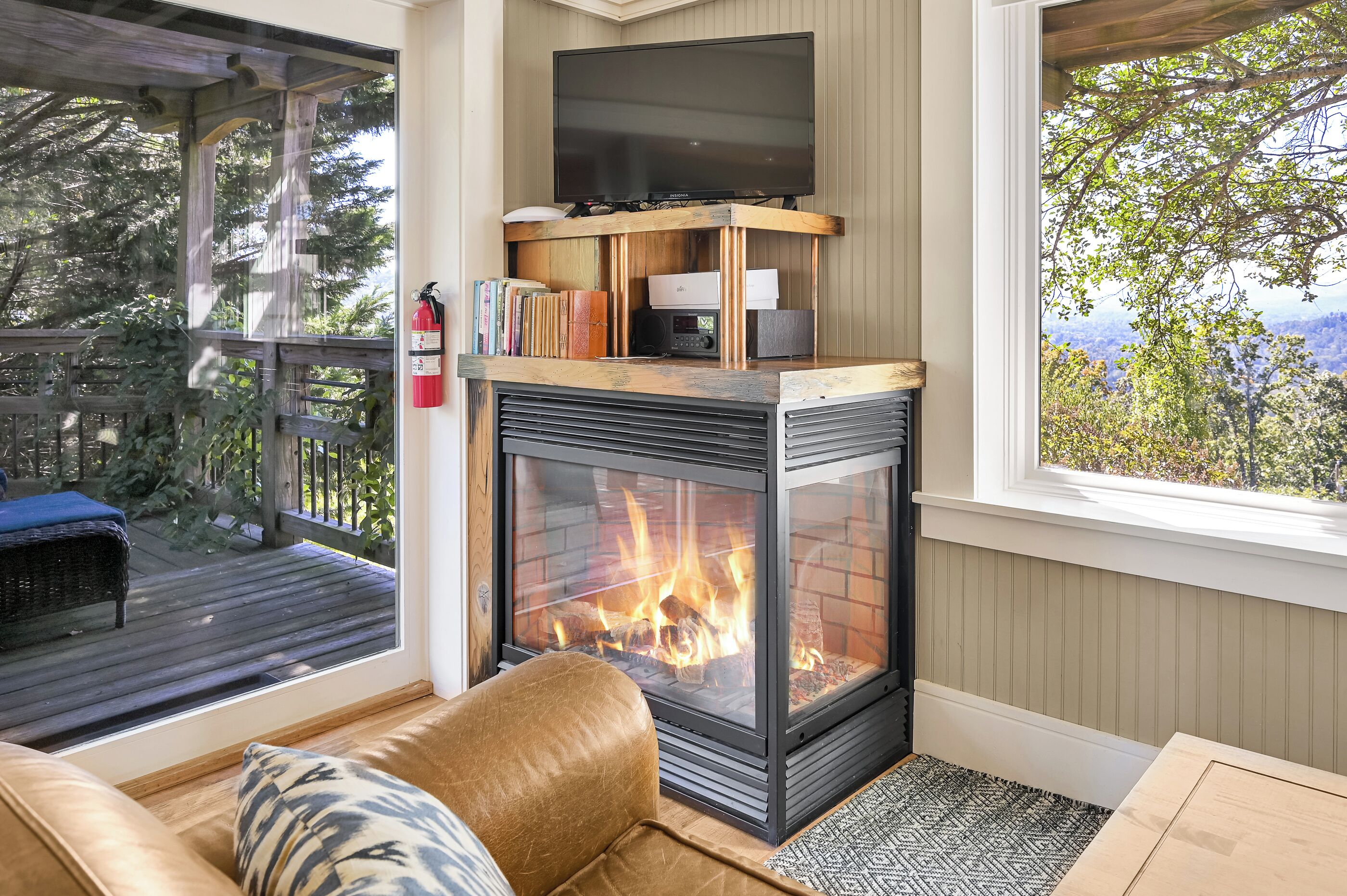 Un salon chaleureux avec une cheminée, un téléviseur posé sur un meuble en bois et une vue sur l’extérieur.
