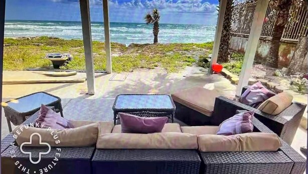 Un área de estar al aire libre cubierta, con mobiliario de mimbre y cojines, con vista a una playa con palmeras y el océano.