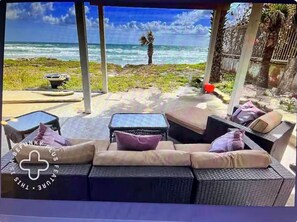 Un área de estar al aire libre cubierta, con mobiliario de mimbre y cojines, con vista a una playa con palmeras y el océano.