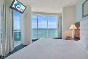 A hotel room with a large bed, a TV mounted on the wall, a nightstand with a lamp, and a view of the sea through large windows. - New HIGH 2BR 20th FLOOR, Direct Oceanfront, Pools! (Myrtle Beach)